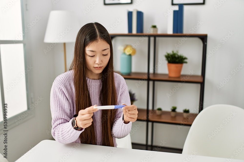 Young chinese girl holding pregnancy test sitting on the table at home. Young chinese girl holding pregnancy test sitting on the table at home.