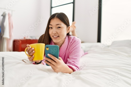 Young chinese girl using smartphone and drinking coffee lying on the bed at bedroom.