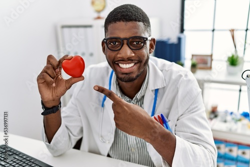 Young african american doctor man wearing doctor uniform holding heart at the clinic smiling happy pointing with hand and finger © Krakenimages.com