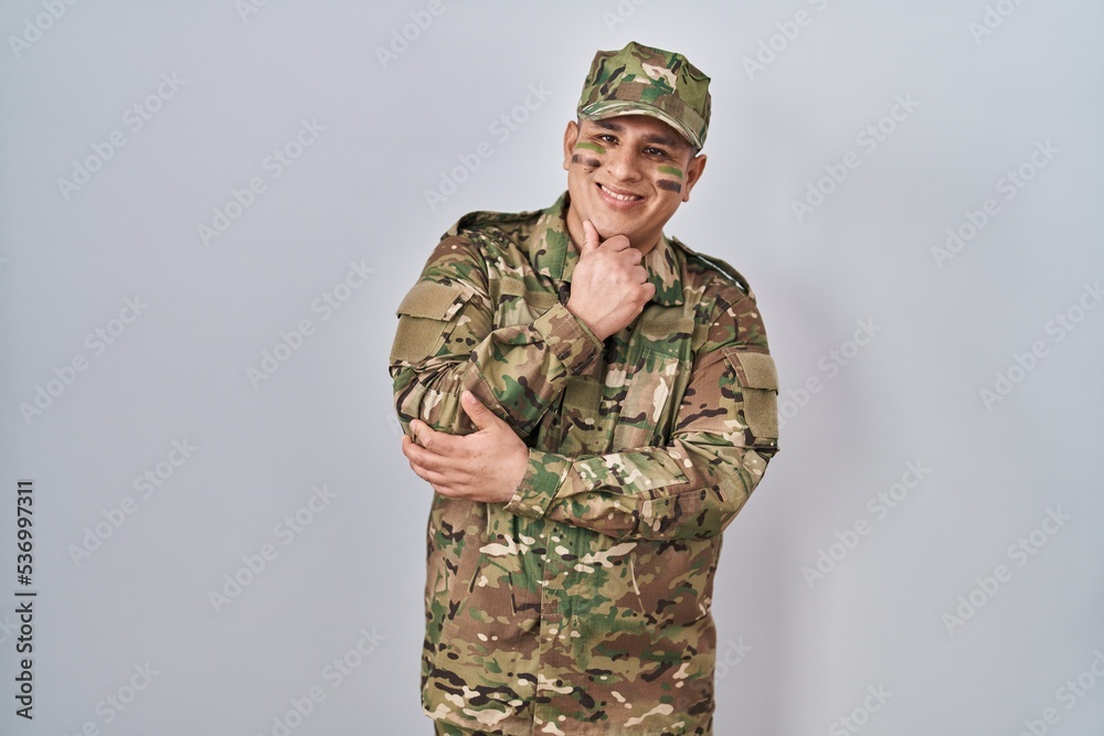 Hispanic young man wearing camouflage army uniform looking confident at the camera smiling with crossed arms and hand raised on chin. thinking positive. Hispanic young man wearing camouflage army uniform looking confident at the camera smiling with crossed arms and hand raised on chin. thinking positive.