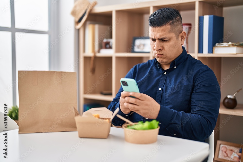 Hispanic young man eating take away food using smartphone skeptic and nervous, frowning upset because of problem. negative person. Hispanic young man eating take away food using smartphone skeptic and nervous, frowning upset because of problem. negative person.