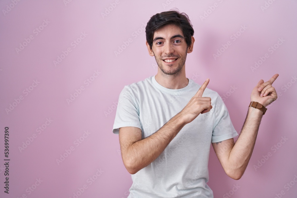 Young hispanic man standing over pink background smiling and looking at the camera pointing with two hands and fingers to the side. Young hispanic man standing over pink background smiling and looking at the camera pointing with two hands and fingers to the side.