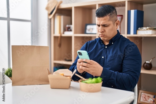 Hispanic young man eating take away food using smartphone skeptic and nervous, frowning upset because of problem. negative person.