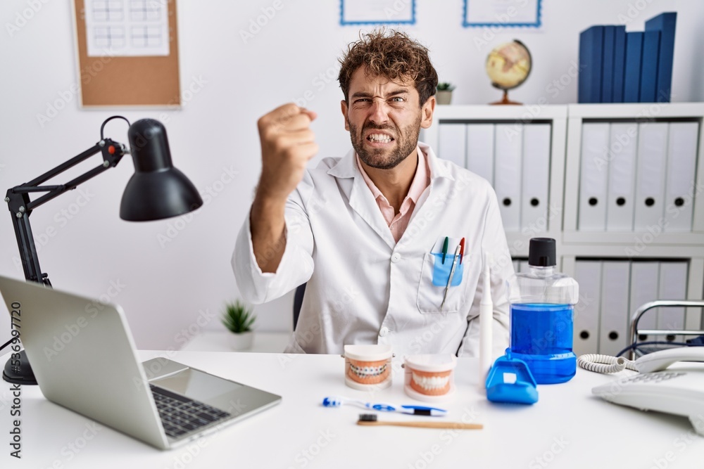 Young hispanic dentist man working at medical clinic angry and mad raising fist frustrated and furious while shouting with anger. rage and aggressive concept. Young hispanic dentist man working at medical clinic angry and mad raising fist frustrated and furious while shouting with anger. rage and aggressive concept.