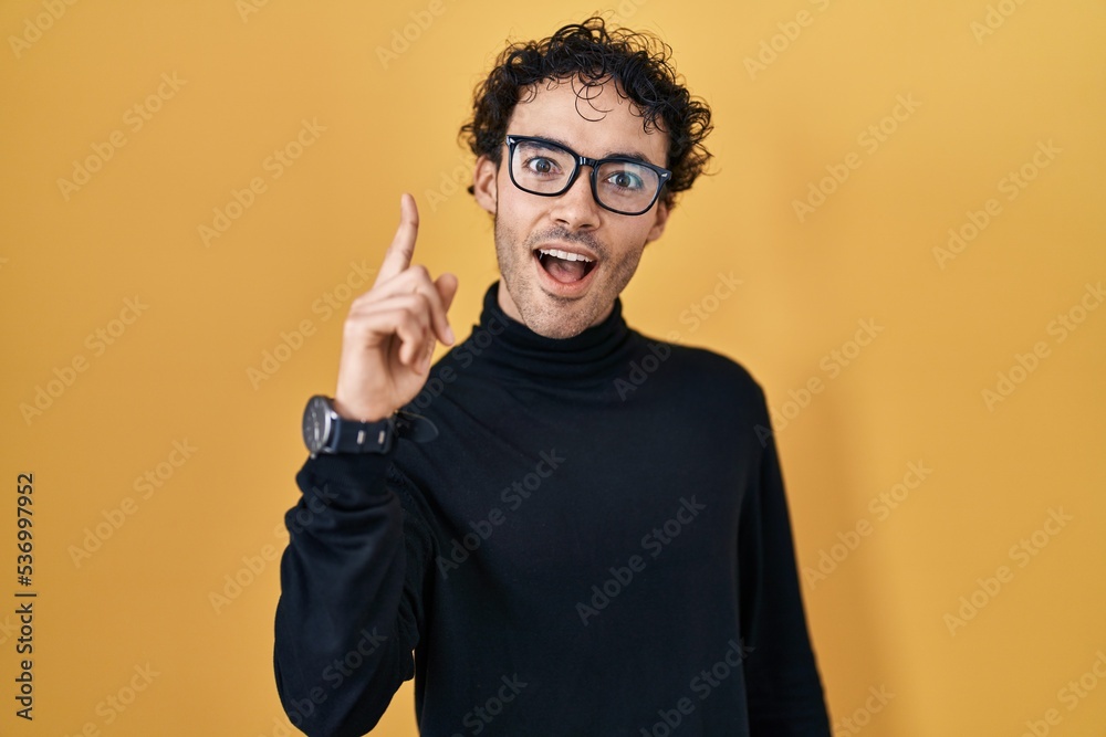 Hispanic man standing over yellow background pointing finger up with successful idea. exited and happy. number one.