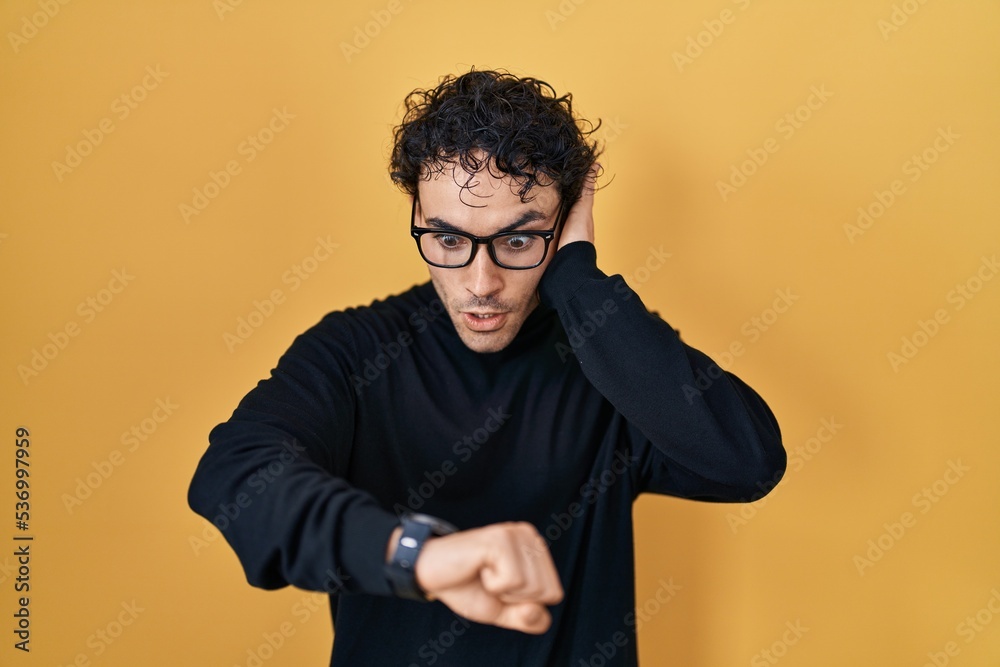 Hispanic man standing over yellow background looking at the watch time worried, afraid of getting late Hispanic man standing over yellow background looking at the watch time worried, afraid of getting late