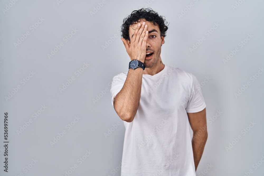 Hispanic man standing over isolated background covering one eye with hand, confident smile on face and surprise emotion.