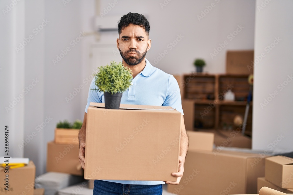 Hispanic man with beard moving to a new home holding box puffing cheeks with funny face. mouth inflated with air, catching air. Hispanic man with beard moving to a new home holding box puffing cheeks with funny face. mouth inflated with air, catching air.