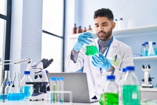 Young hispanic man wearing scientist uniform using laptop holding test tube at laboratory