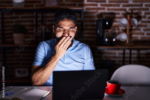 Hispanic man with beard using laptop at night bored yawning tired covering mouth with hand. restless and sleepiness.
