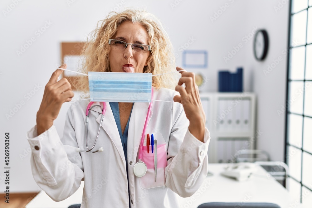 Middle age blonde woman wearing doctor uniform and medical mask sticking tongue out happy with funny expression. Middle age blonde woman wearing doctor uniform and medical mask sticking tongue out happy with funny expression.