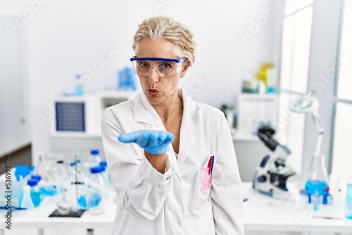Middle age blonde woman working at scientist laboratory looking at the camera blowing a kiss with hand on air being lovely and sexy. love expression.
