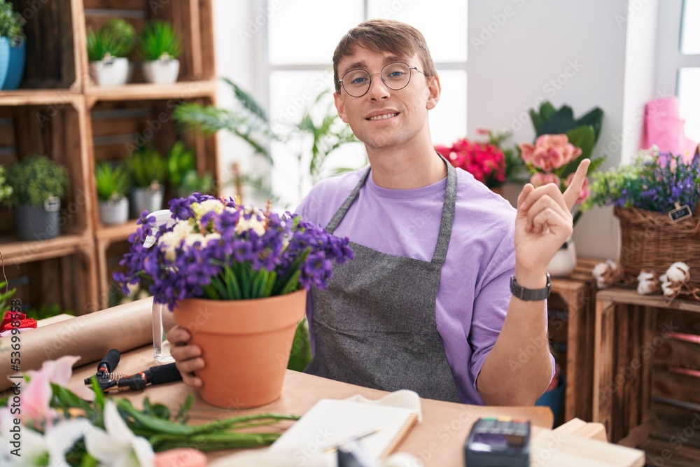 Caucasian blond man working at florist shop with a big smile on face, pointing with hand finger to the side looking at the camera.