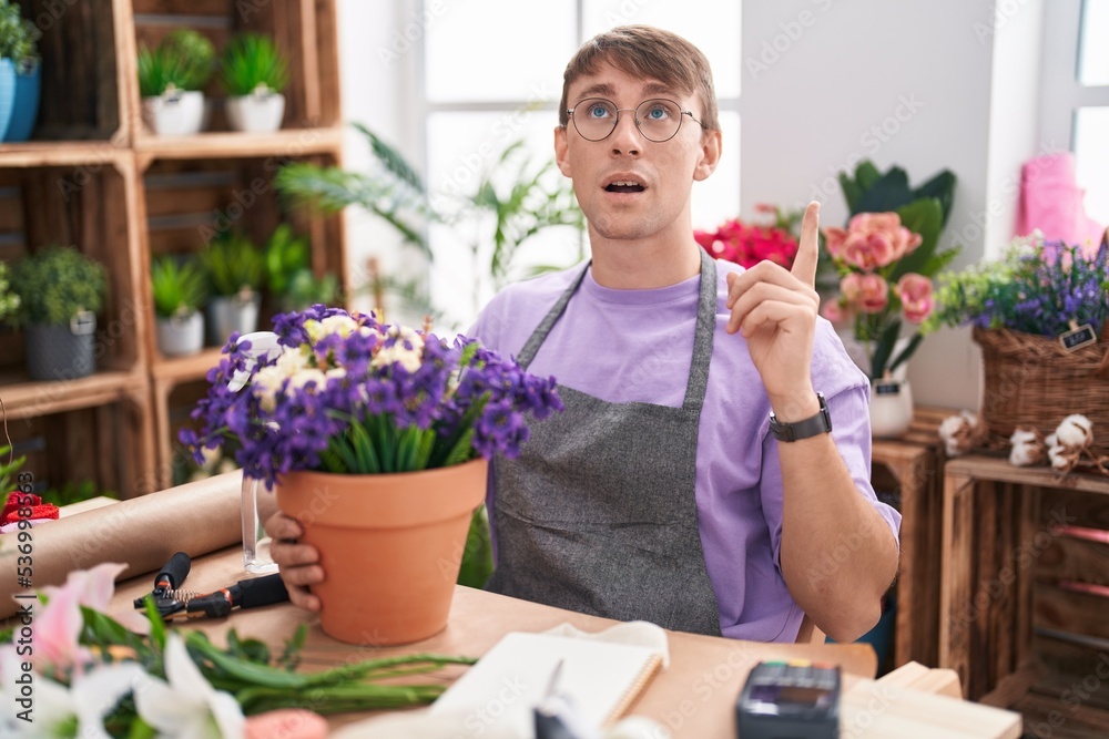 Caucasian blond man working at florist shop amazed and surprised looking up and pointing with fingers and raised arms.