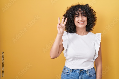 Young middle east woman standing over yellow background showing and pointing up with fingers number three while smiling confident and happy.