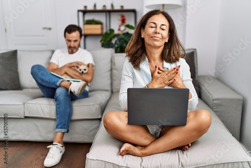 Hispanic middle age couple at home, woman using laptop smiling with hands on chest with closed eyes and grateful gesture on face. health concept.