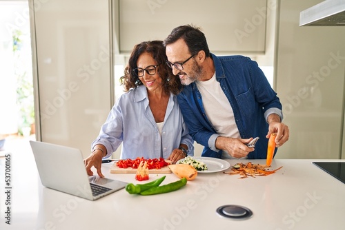 Middle age hispanic couple cooking and using laptop at kitchen