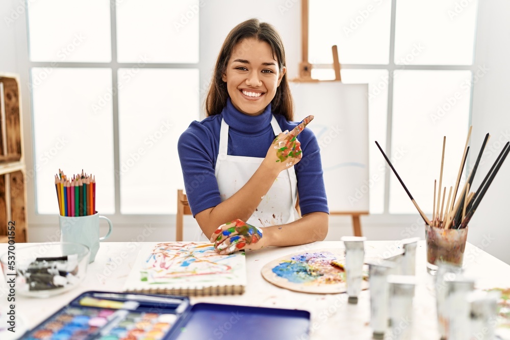 Young brunette woman at art studio with painted hands cheerful with a smile of face pointing with hand and finger up to the side with happy and natural expression on face