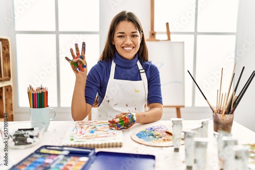 Young brunette woman at art studio with painted hands showing and pointing up with fingers number four while smiling confident and happy.