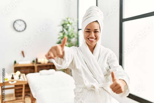 Young brunette woman wearing towel and bathrobe standing at beauty center approving doing positive gesture with hand, thumbs up smiling and happy for success. winner gesture.