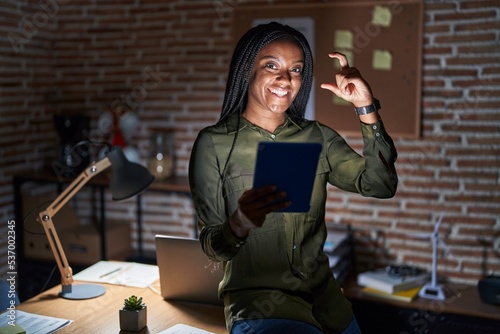 Young african american with braids working at the office at night smiling and confident gesturing with hand doing small size sign with fingers looking and the camera. measure concept.