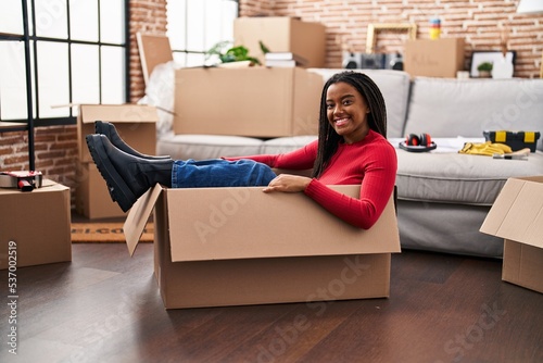 Young african american with braids moving to a new home inside of a cardboard box looking positive and happy standing and smiling with a confident smile showing teeth © Krakenimages.com