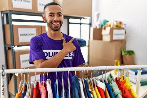 African american man wearing volunteer t shirt at donations stand cheerful with a smile of face pointing with hand and finger up to the side with happy and natural expression on face