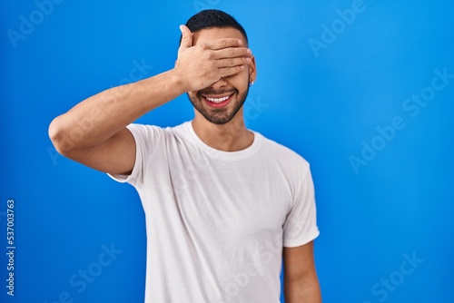 Young hispanic man standing over blue background smiling and laughing with hand on face covering eyes for surprise. blind concept.