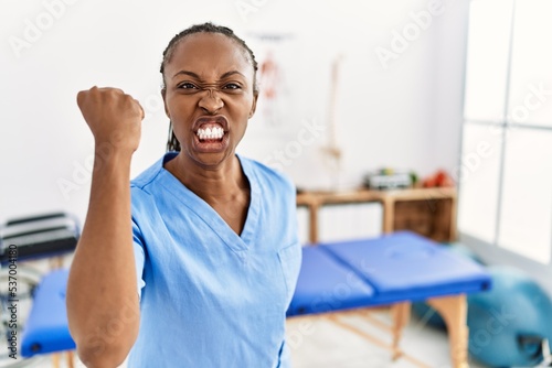Black woman with braids working at pain recovery clinic angry and mad raising fist frustrated and furious while shouting with anger. rage and aggressive concept.