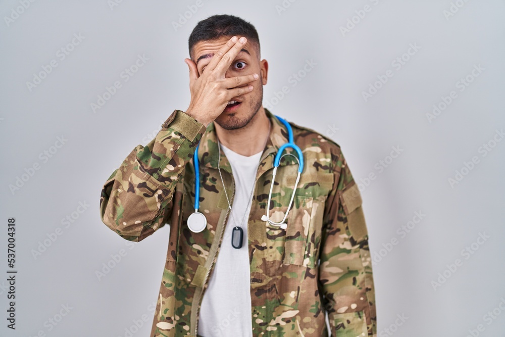 Young hispanic doctor wearing camouflage army uniform peeking in shock covering face and eyes with hand, looking through fingers afraid