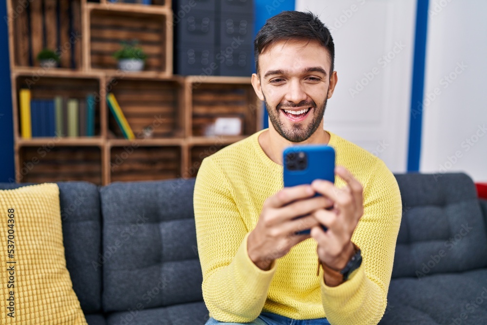 Young hispanic man using smartphone sitting on sofa at home Young hispanic man using smartphone sitting on sofa at home