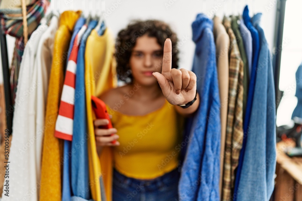 Young hispanic woman searching clothes on clothing rack using smartphone pointing with finger up and angry expression, showing no gesture