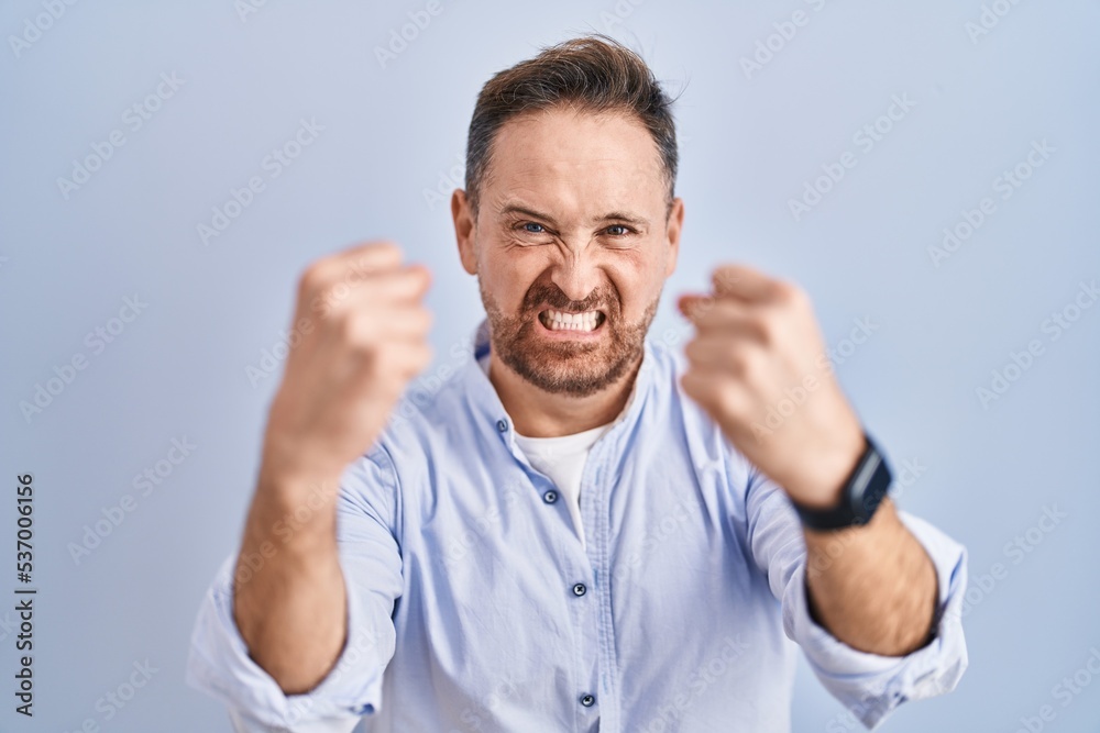 Middle age caucasian man standing over blue background angry and mad raising fists frustrated and furious while shouting with anger. rage and aggressive concept. Middle age caucasian man standing over blue background angry and mad raising fists frustrated and furious while shouting with anger. rage and aggressive concept.