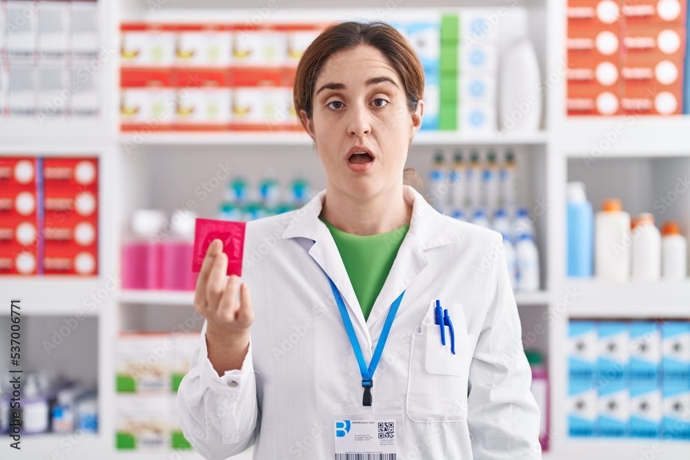 Brunette woman working at pharmacy drugstore holding condom scared and amazed with open mouth for surprise, disbelief face