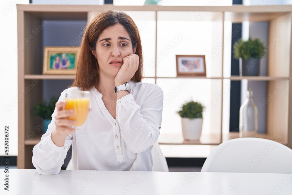 Brunette woman drinking glass of orange juice looking stressed and nervous with hands on mouth biting nails. anxiety problem. Brunette woman drinking glass of orange juice looking stressed and nervous with hands on mouth biting nails. anxiety problem.