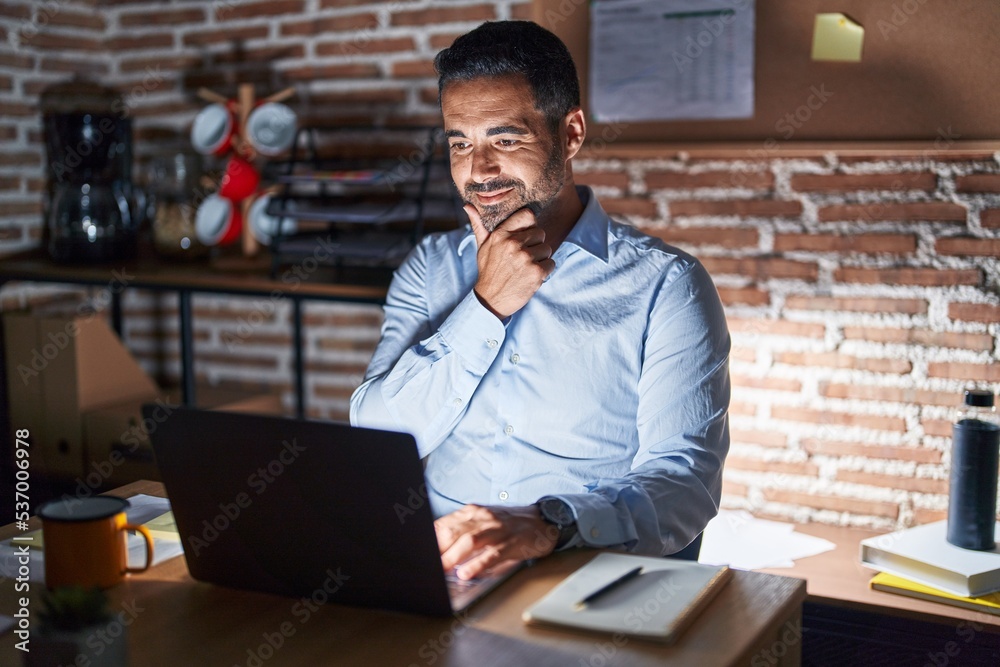 Hispanic man with beard working at the office at night looking confident at the camera smiling with crossed arms and hand raised on chin. thinking positive. Hispanic man with beard working at the office at night looking confident at the camera smiling with crossed arms and hand raised on chin. thinking positive.