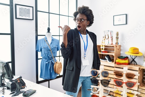 African young woman working as manager at retail boutique surprised pointing with hand finger to the side, open mouth amazed expression.