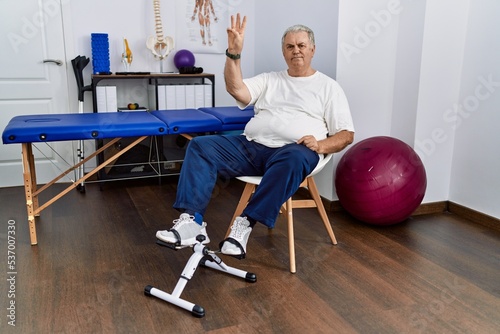 Senior caucasian man at physiotherapy clinic using pedal exerciser showing and pointing up with fingers number three while smiling confident and happy.