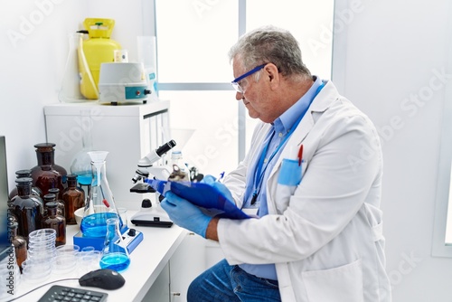 Middle age grey-haired man wearing scientist uniform writing on clipboard at laboratory