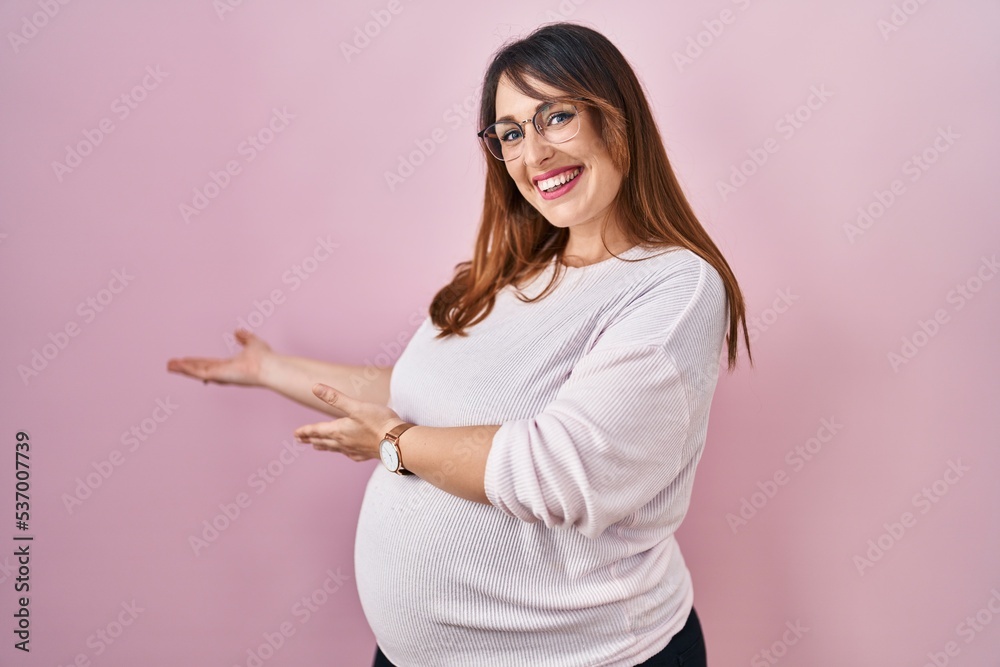 Pregnant woman standing over pink background inviting to enter smiling natural with open hand Pregnant woman standing over pink background inviting to enter smiling natural with open hand