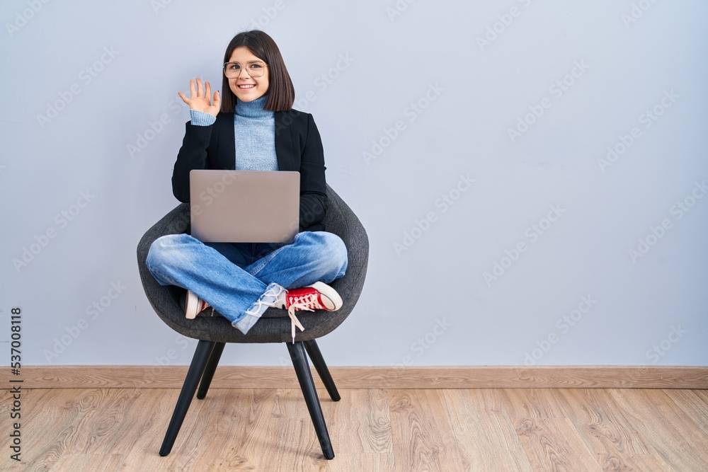 Young hispanic woman sitting on chair using computer laptop waiving saying hello happy and smiling, friendly welcome gesture Young hispanic woman sitting on chair using computer laptop waiving saying hello happy and smiling, friendly welcome gesture