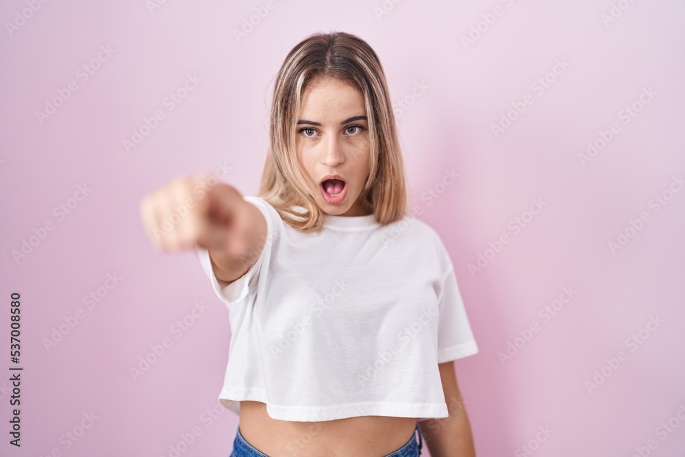 Young blonde woman standing over pink background pointing with finger surprised ahead, open mouth amazed expression, something on the front Young blonde woman standing over pink background pointing with finger surprised ahead, open mouth amazed expression, something on the front