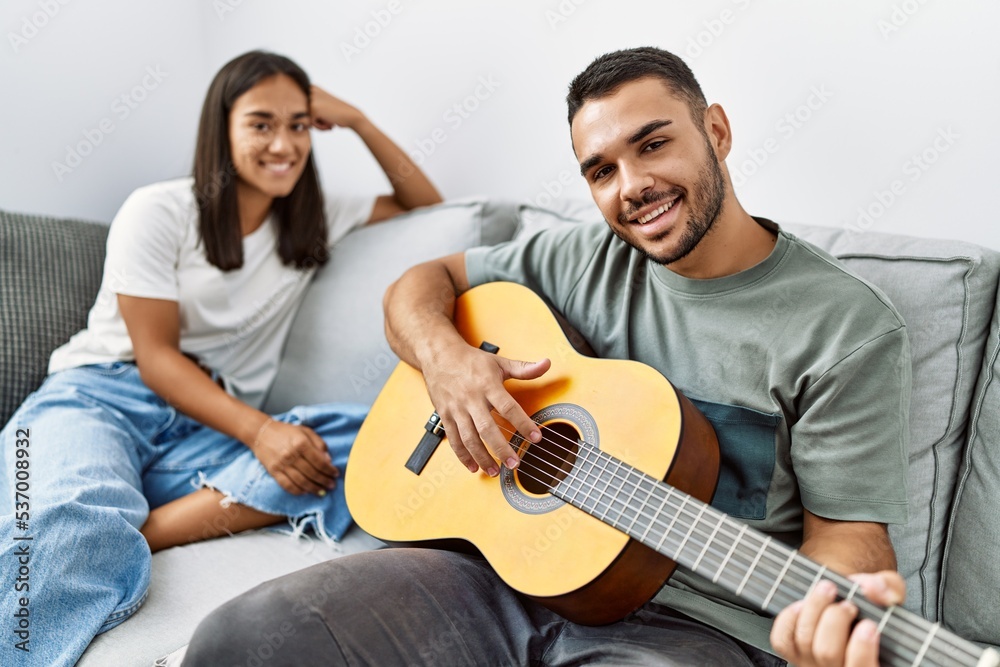 Fototapeta premium Young latin couple playing classical guitar sitting on the sofa at home.