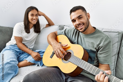 Young latin couple playing classical guitar sitting on the sofa at home.