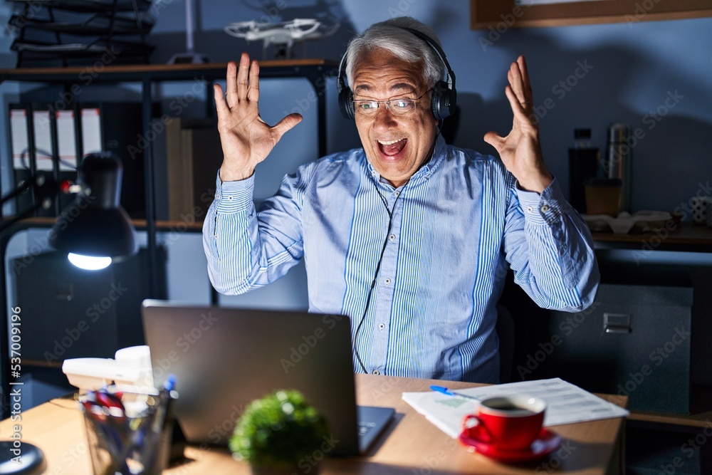 Fototapeta premium Hispanic senior man wearing call center agent headset at night celebrating crazy and amazed for success with arms raised and open eyes screaming excited. winner concept