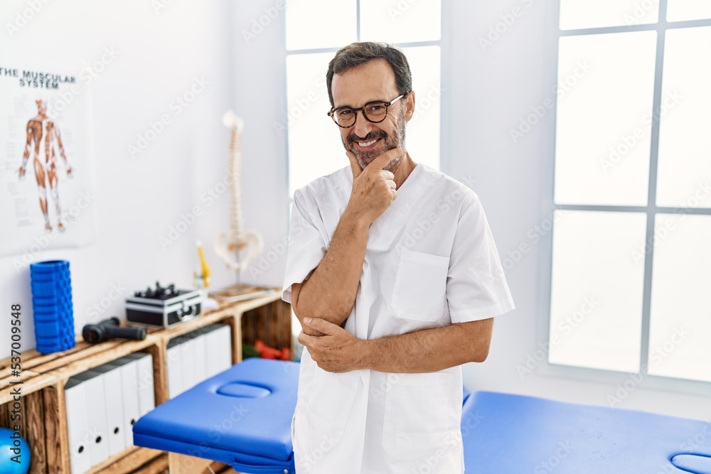 Middle age man with beard working at pain recovery clinic looking confident at the camera smiling with crossed arms and hand raised on chin. thinking positive. Middle age man with beard working at pain recovery clinic looking confident at the camera smiling with crossed arms and hand raised on chin. thinking positive.