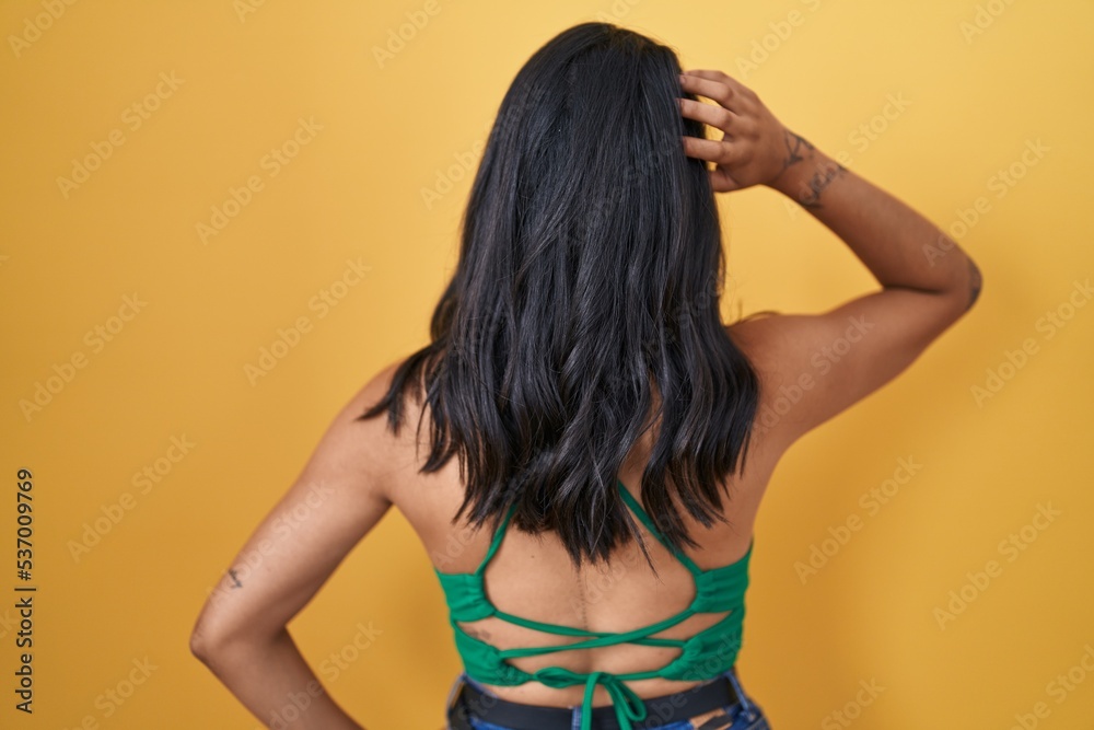 Brunette woman standing over yellow background backwards thinking about doubt with hand on head Brunette woman standing over yellow background backwards thinking about doubt with hand on head