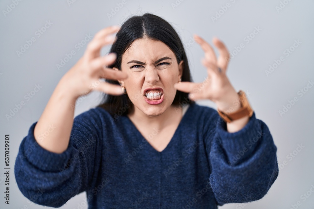 Young brunette woman standing over isolated background shouting frustrated with rage, hands trying to strangle, yelling mad Young brunette woman standing over isolated background shouting frustrated with rage, hands trying to strangle, yelling mad
