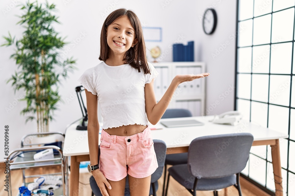 Young hispanic girl standing at pediatrician clinic smiling cheerful presenting and pointing with palm of hand looking at the camera. Young hispanic girl standing at pediatrician clinic smiling cheerful presenting and pointing with palm of hand looking at the camera.