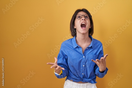 Young girl standing over yellow background crazy and mad shouting and yelling with aggressive expression and arms raised. frustration concept.